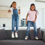 Excited girl kids jumping on living room sofa furniture at home for fun, energy and play games. Portrait crazy, happy and wild young children adhd, action and funny laughing bounce on lounge couch